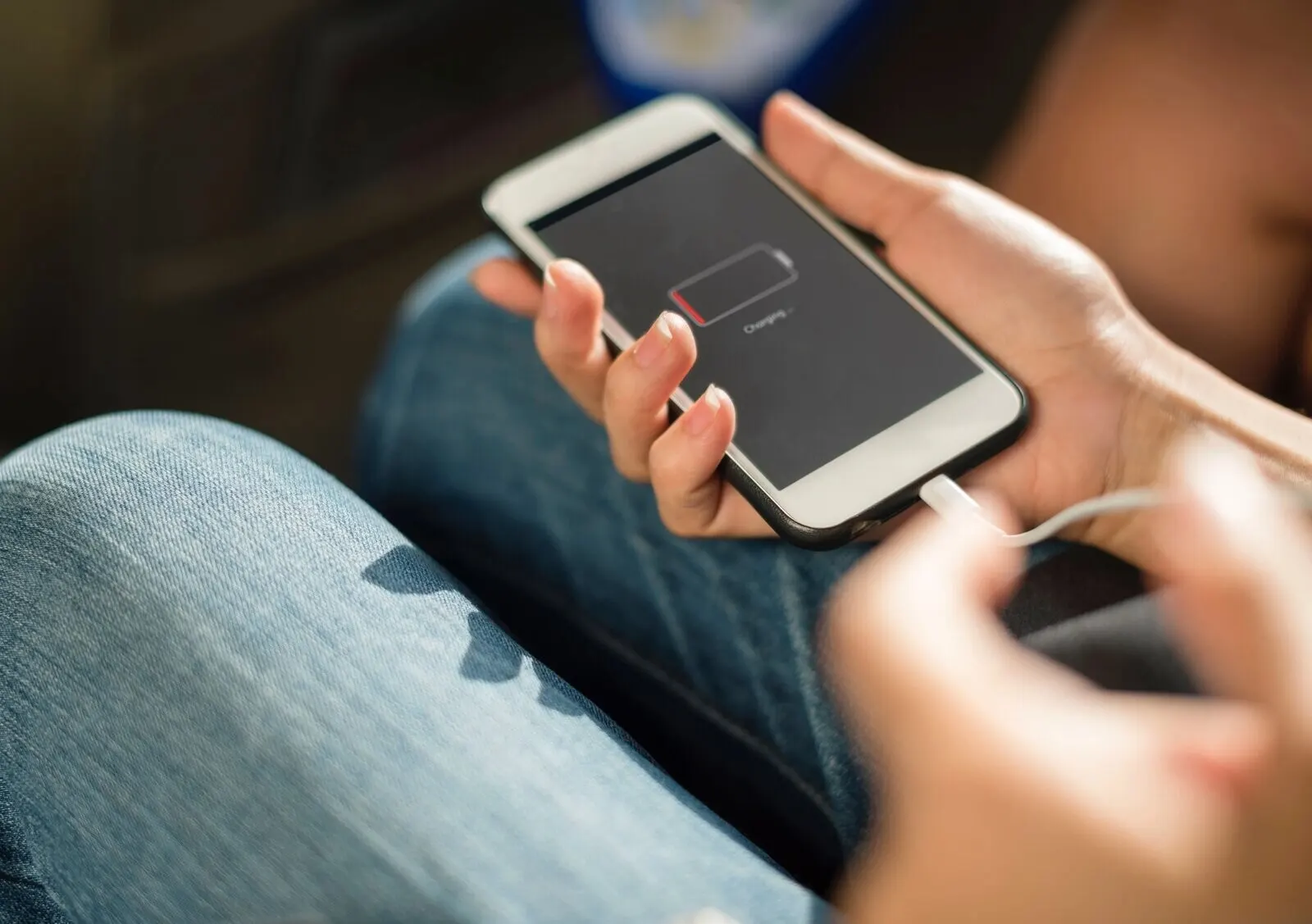 Hands holding a white iPhone that shows an empty-battery icon and the word "Charging", with a short white cable plugged in, over a pair of jeans