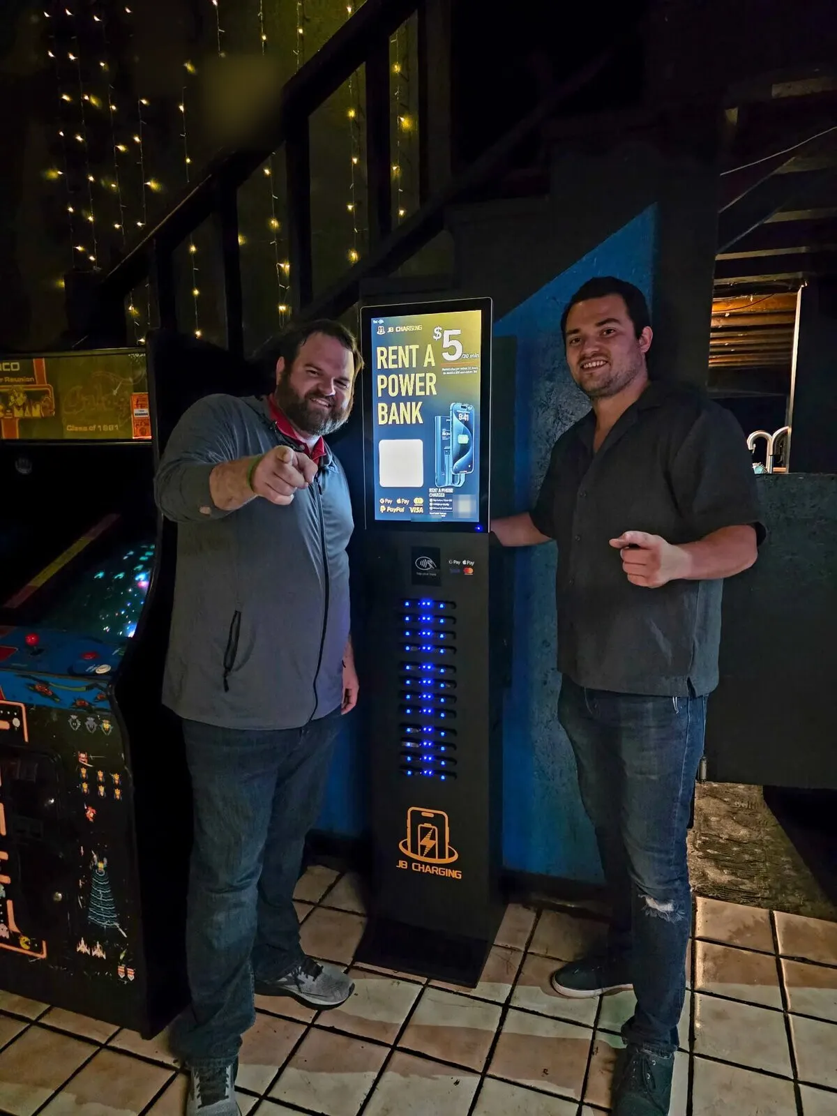 Two men pointing toward the camera beside a JB Charging J24 Pro tower whose screen reads Rent a Power Bank, $5 per 30 minutes, next to retro arcade cabinets in a dim room with string lights