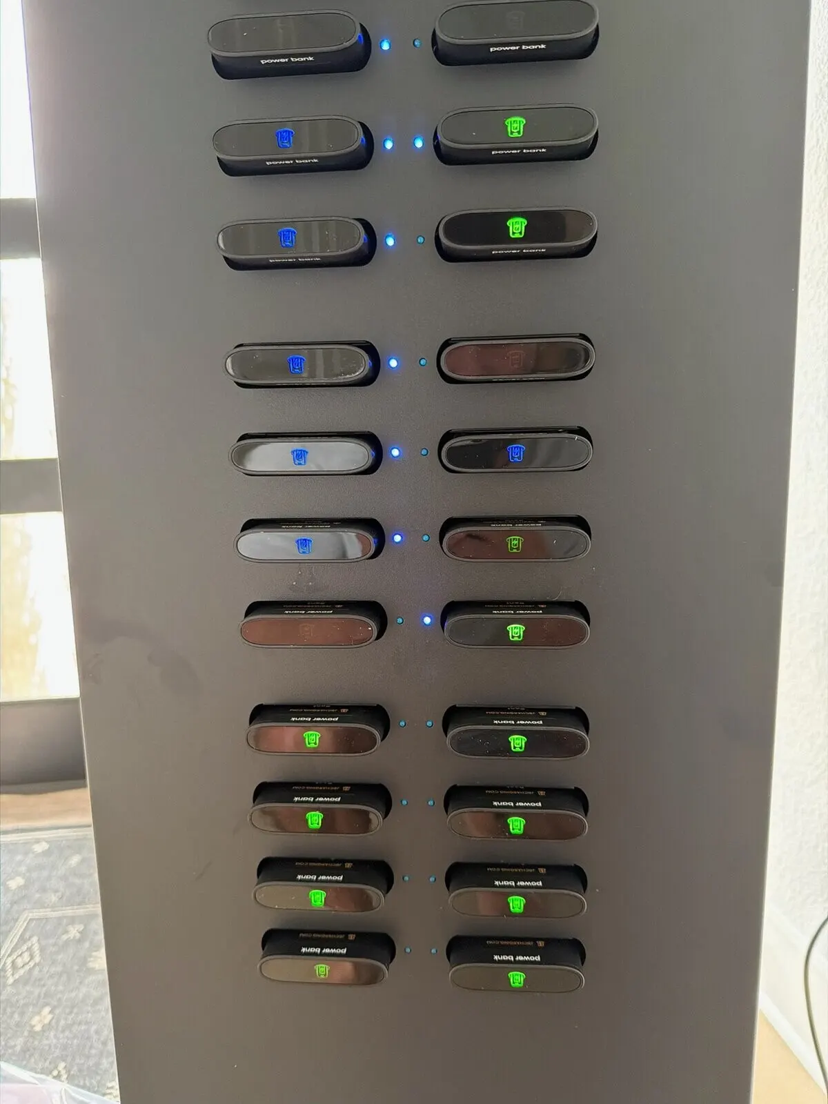Close-up of a J24 Pro tower's slot column, with black power banks docked in two columns, green and blue charge icons lit on the banks and small blue status lights between the columns
