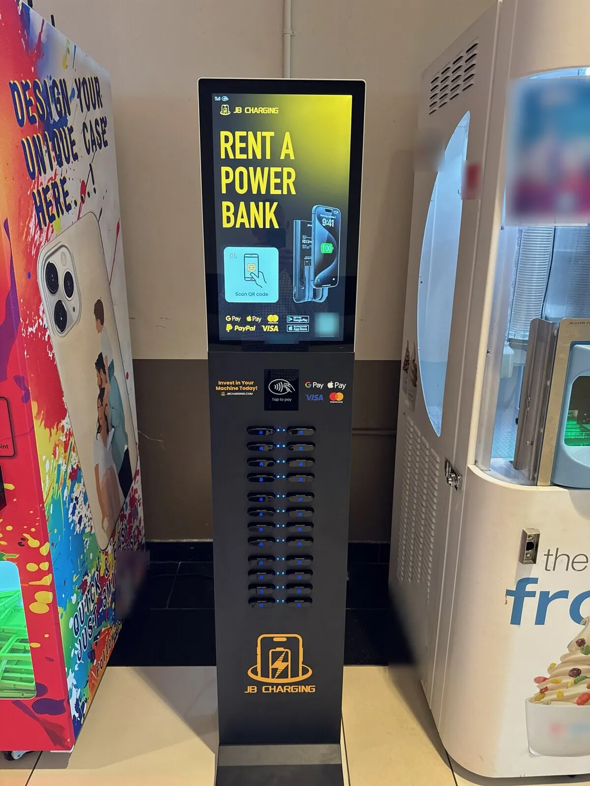 A JB Charging tower station standing between a colorful self-service phone case machine and a frozen yogurt vending machine