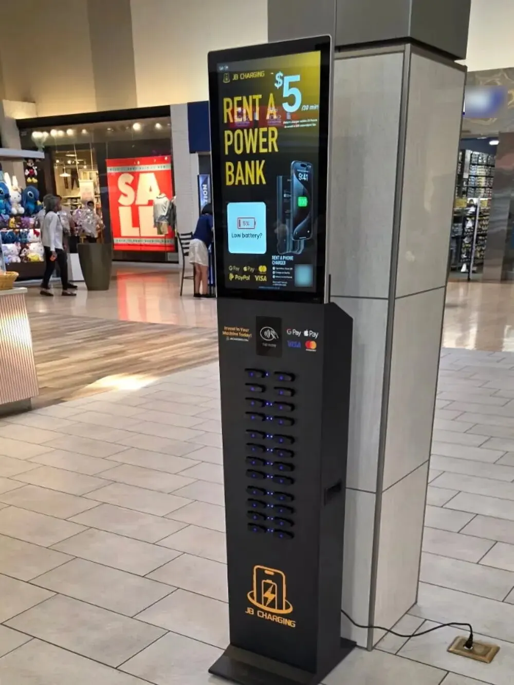 A JB Charging J24 Pro floor tower beside a pillar in a shopping-mall concourse, its screen advertising power bank rentals at $5 per 30 minutes