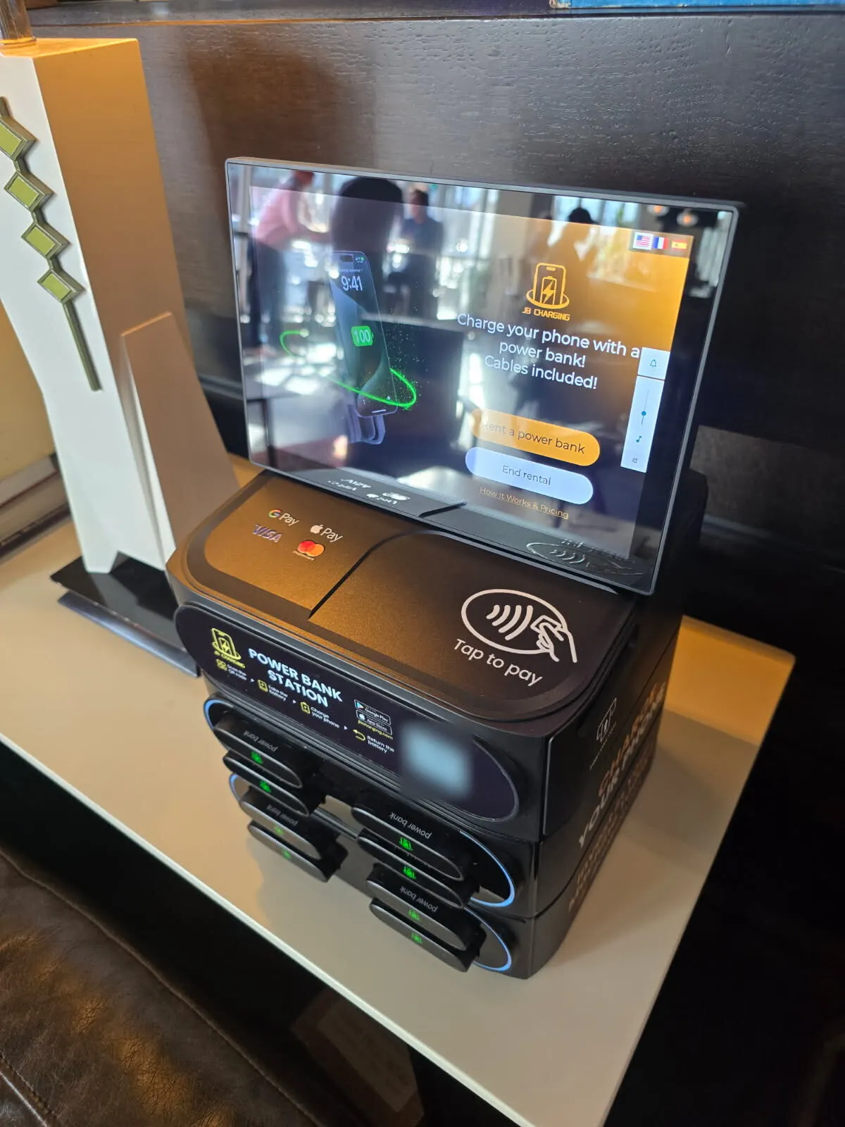 J8 power bank station on a white shelf above a leather seat, its screen showing "Rent a power bank" and "End rental" buttons over the tap-to-pay pad and QR code