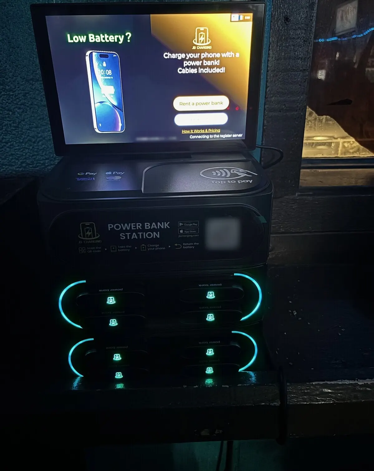 A J8 Pro station in a dim room under a lit sign, its screen showing a "Low Battery?" ad and a "Rent a power bank" button, with small status text near the bottom edge reading "Connecting to the register server"
