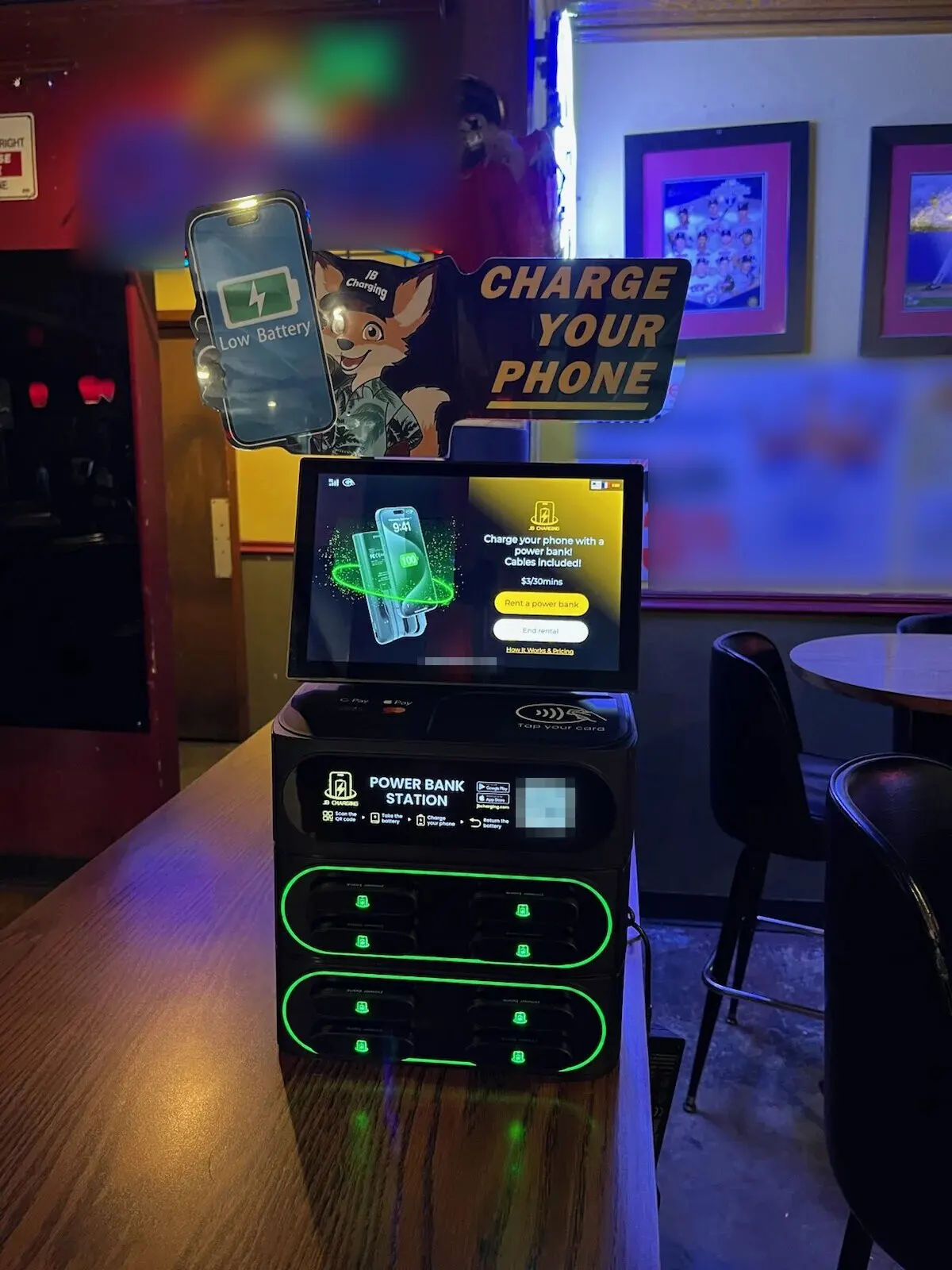 A JB Charging J8 Pro with a "Charge Your Phone" fox header at the end of a wooden bar counter, its screen showing $3 per 30 minutes, with a neon beer sign and framed photos on the wall behind it