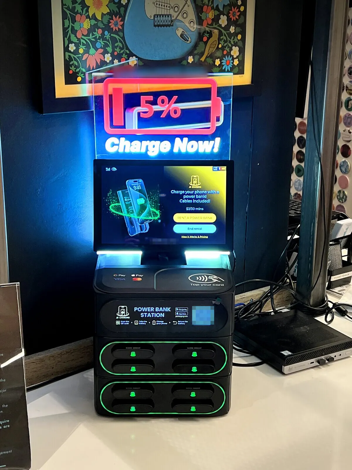 J8 power bank station on a white front-desk counter, with a lit "5% Charge Now" sign above a touchscreen showing $3 per 30 minutes and slots lit green
