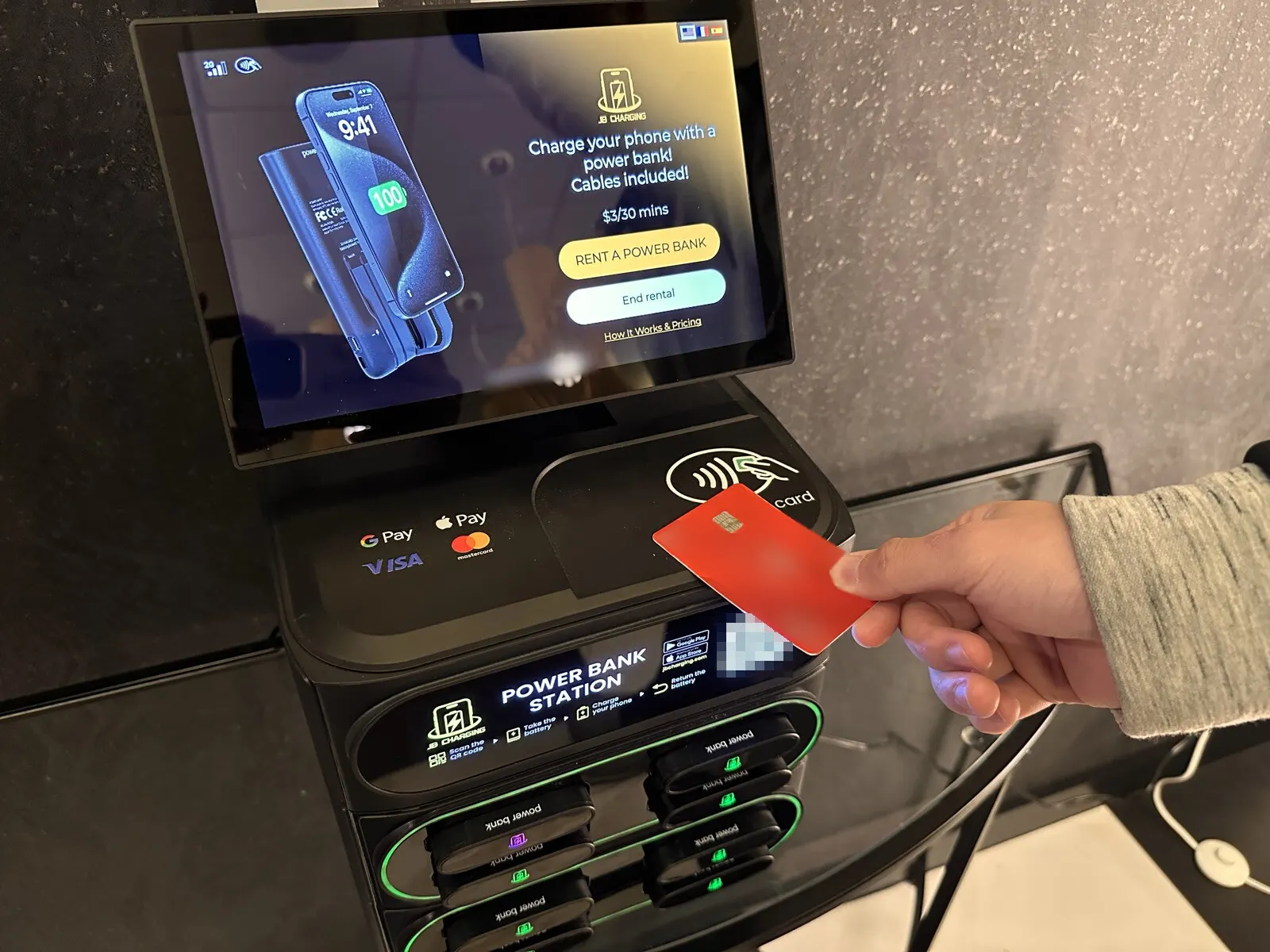 A hand holding a red contactless card to the reader of a JB Charging station whose screen shows $3 per 30 minutes and a Rent a Power Bank button