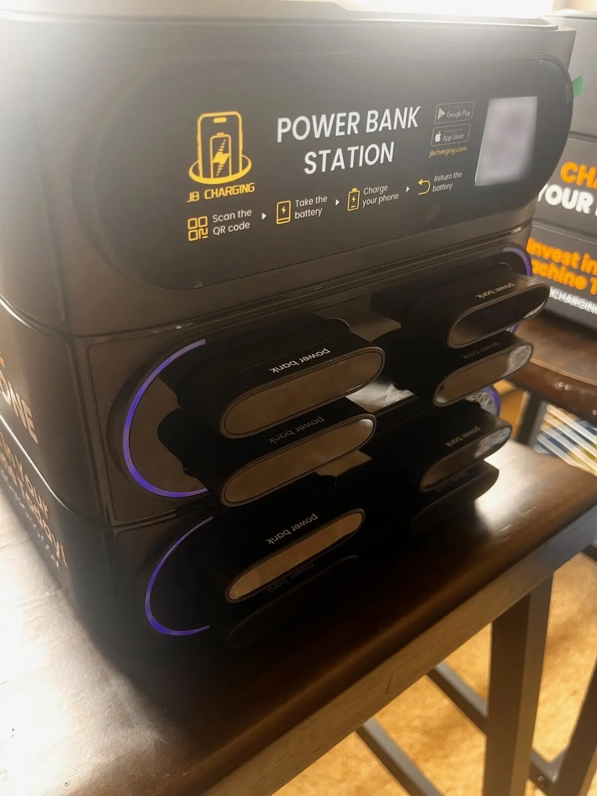 Three-quarter close-up of a JB Charging countertop station's front panel showing the steps Scan the QR code, Take the battery, Charge your phone and Return the battery, above docked power banks in purple-lit slots