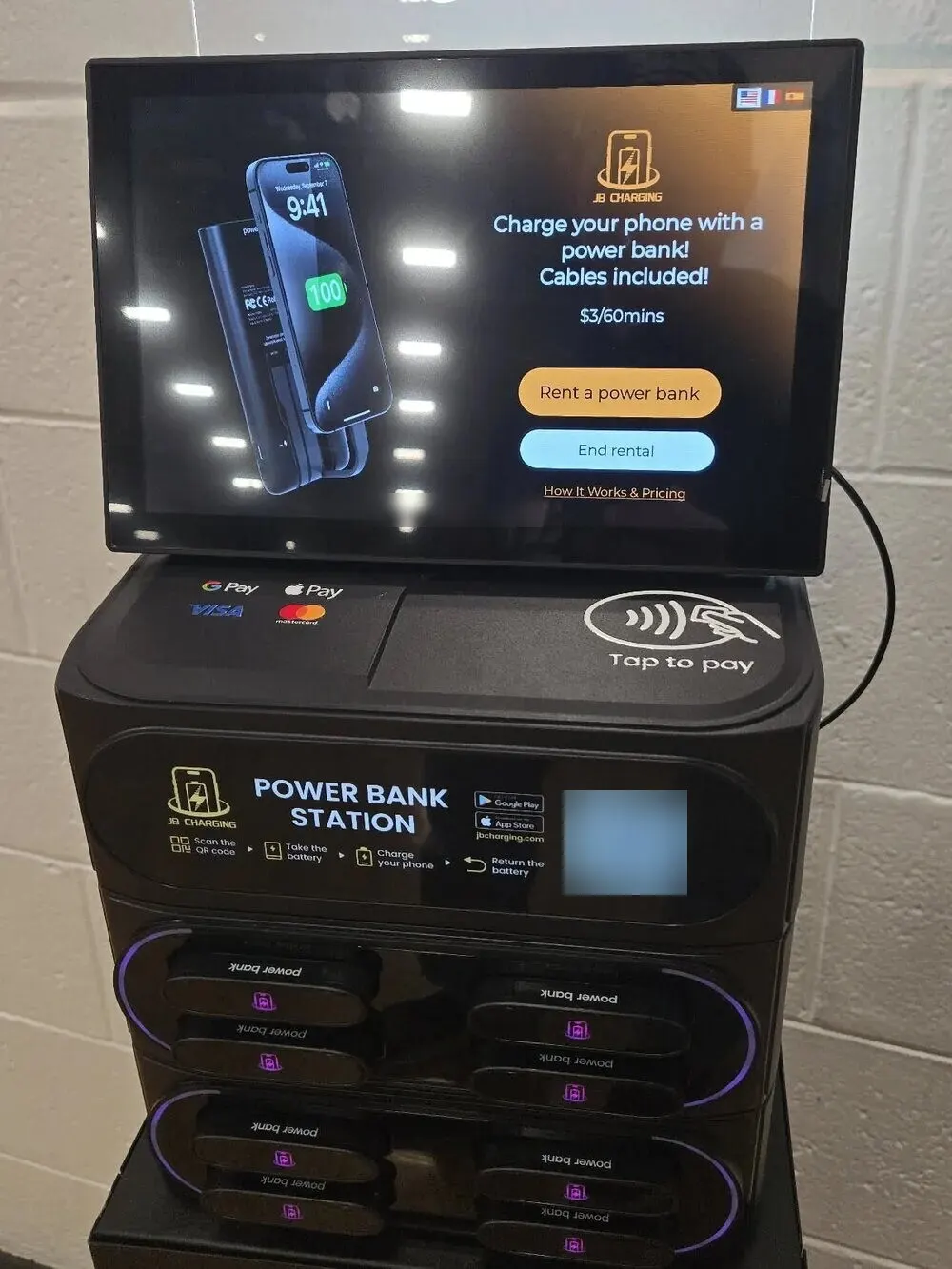 J8 power bank station against a white cinderblock wall, its touchscreen showing $3 per 60 minutes above a "Rent a power bank" button, with a tap-to-pay pad below and eight power banks in purple-lit slots