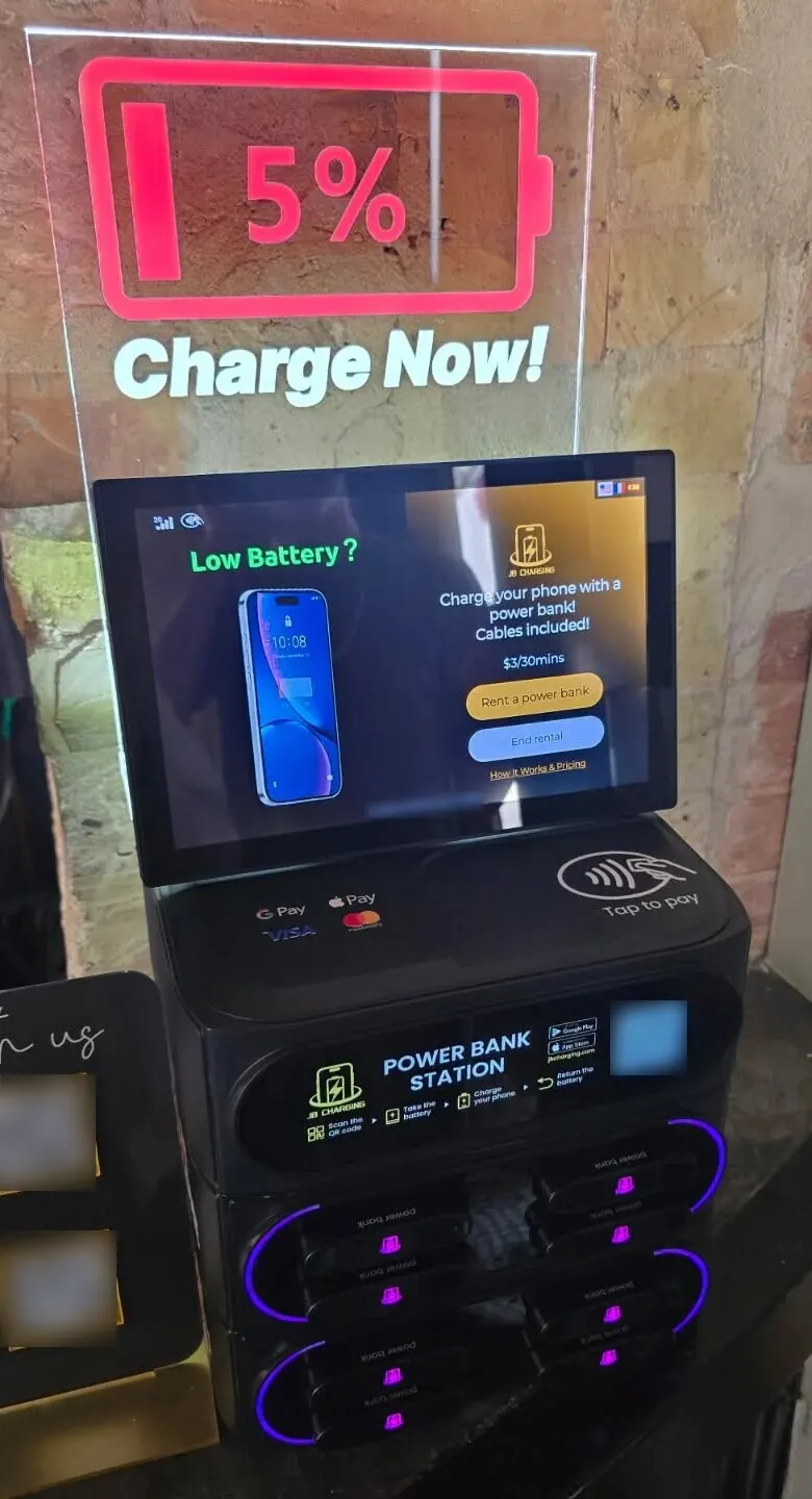 J8 Pro power bank station under a lit "5% Charge Now" sign, its screen showing $3/30mins above Rent a power bank and End rental buttons, against a brick wall