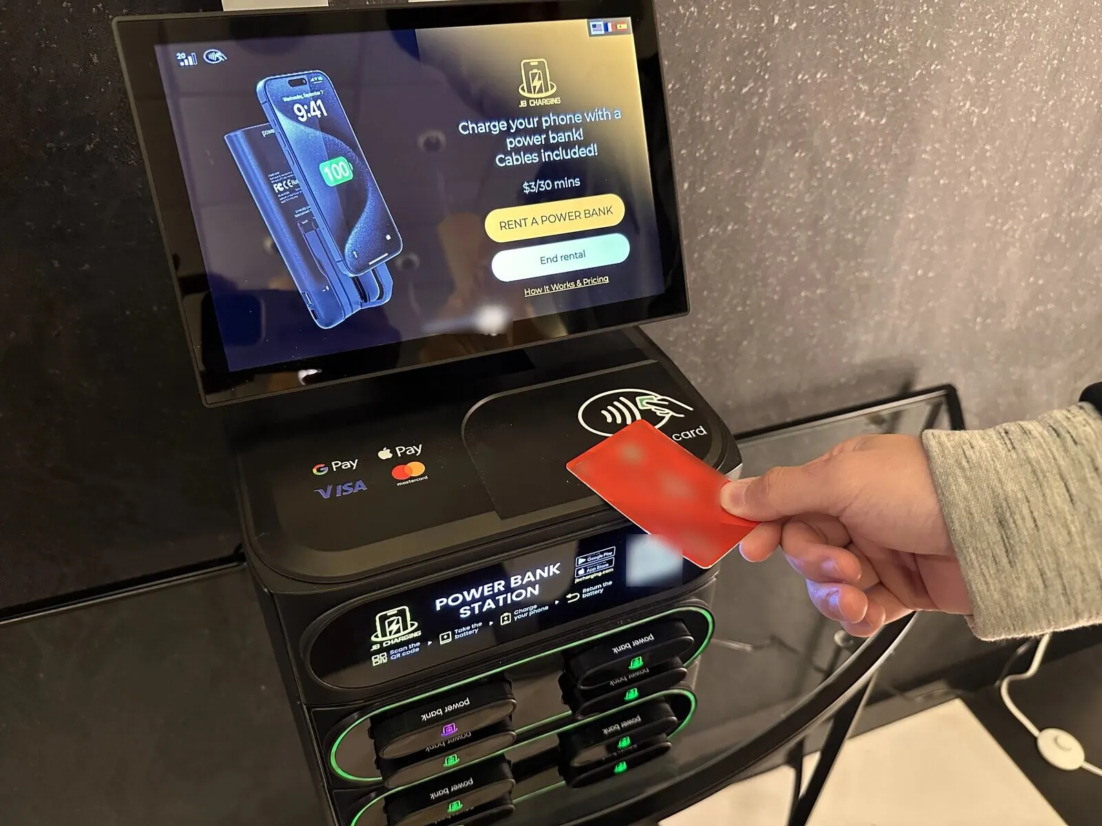 A hand taps a red contactless card on the reader of a J8 power bank station; the screen shows $3 per 30 minutes and a Rent a Power Bank button above power bank slots, most lit green