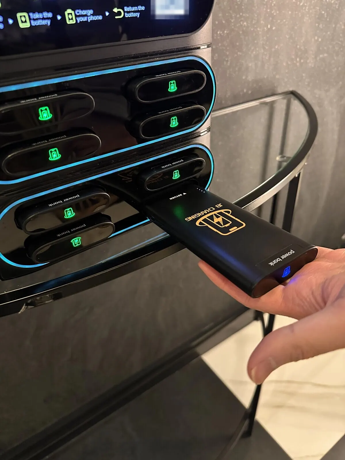 A hand holding a black JB Charging power bank half inside a slot of a countertop station, beside docked banks with glowing green charge icons, on a glass side table