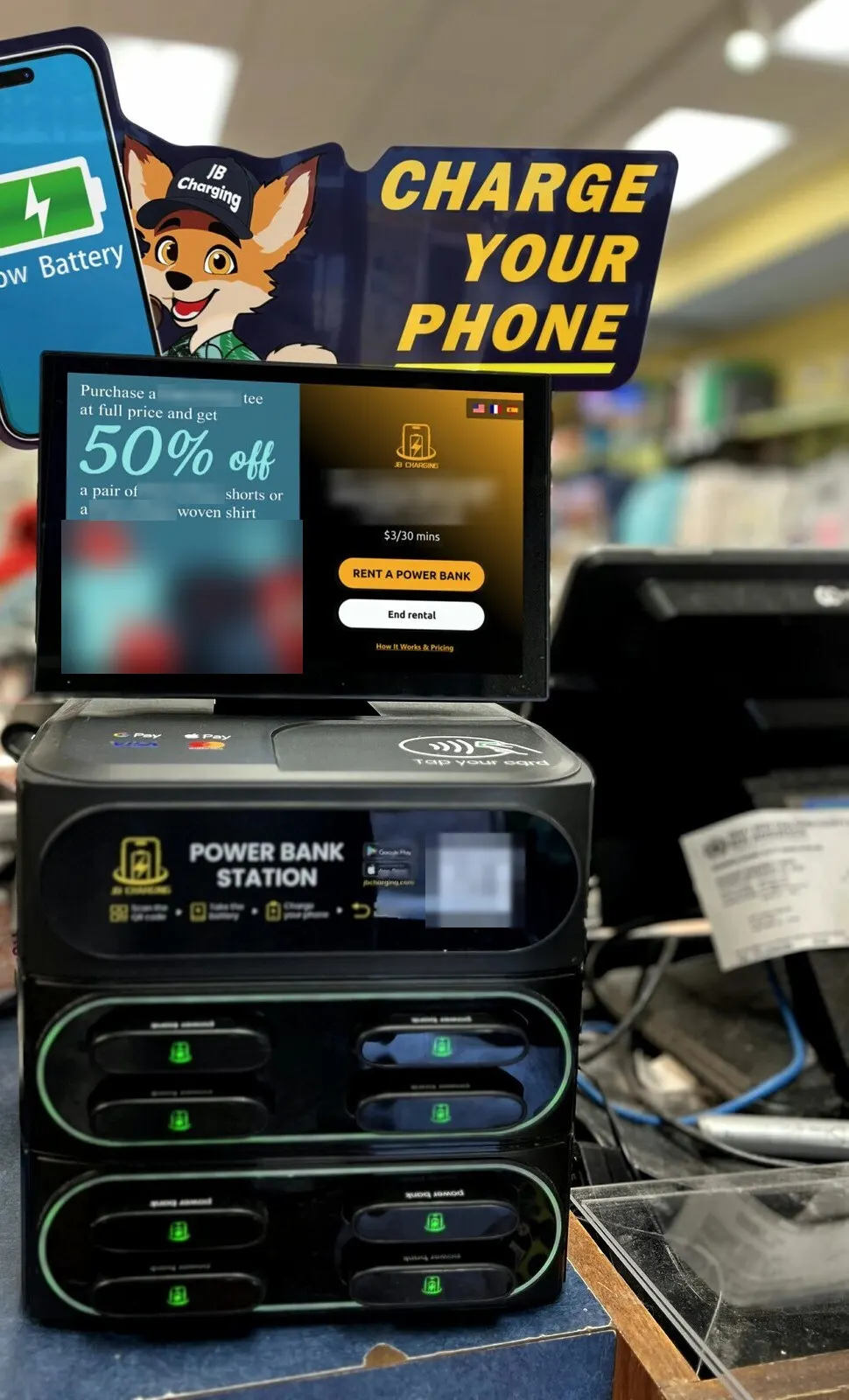 An eight-slot JB Charging station on a shop counter under a Charge Your Phone header sign, its screen split between a clothing promotion and a Rent a Power Bank button