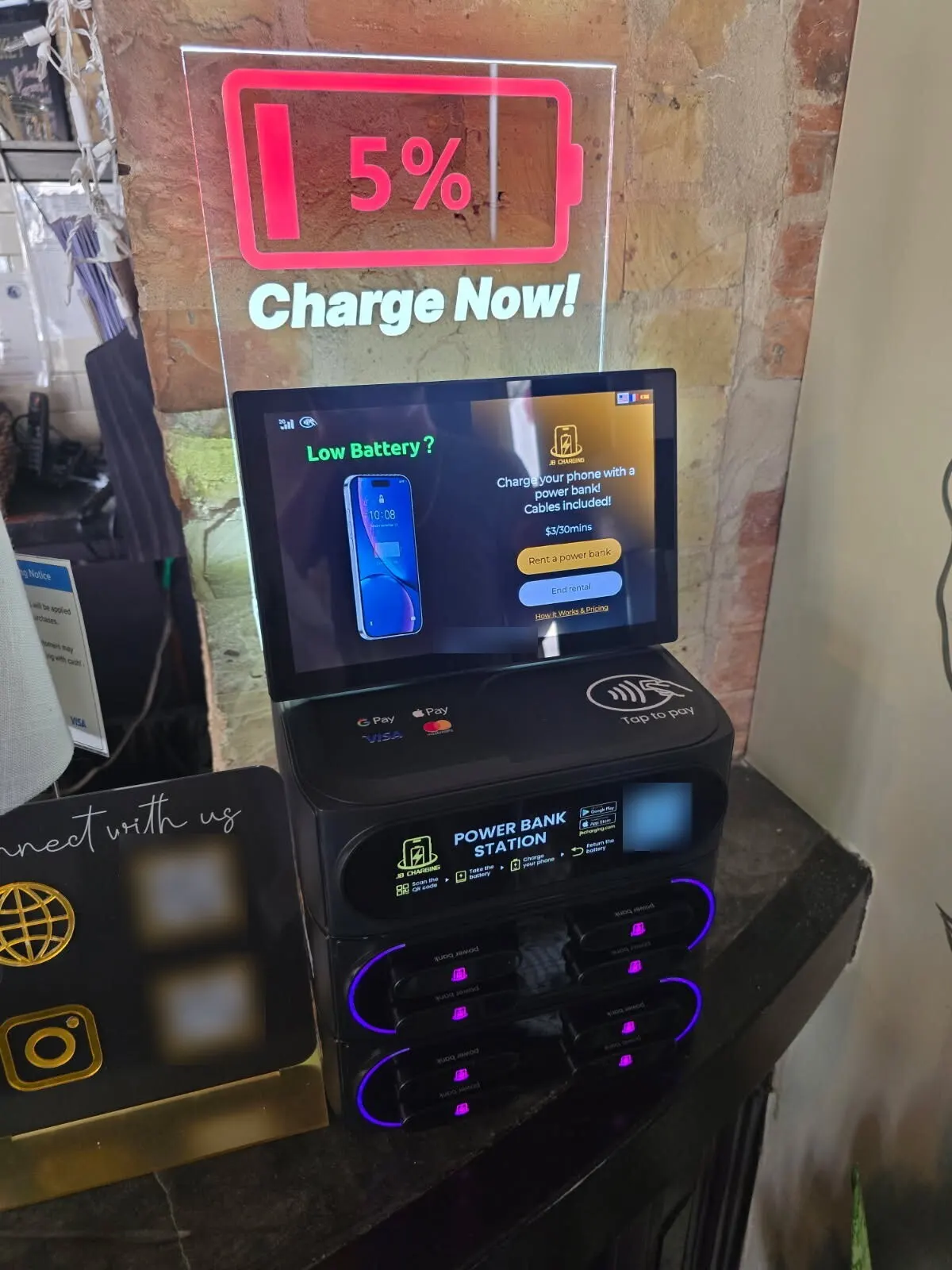 A J8 Pro power bank station on a dark stone counter against a brick wall, under a lit acrylic sign showing a 5% battery and the words "Charge Now," with a tap-to-pay pad on top and eight purple-ringed slots