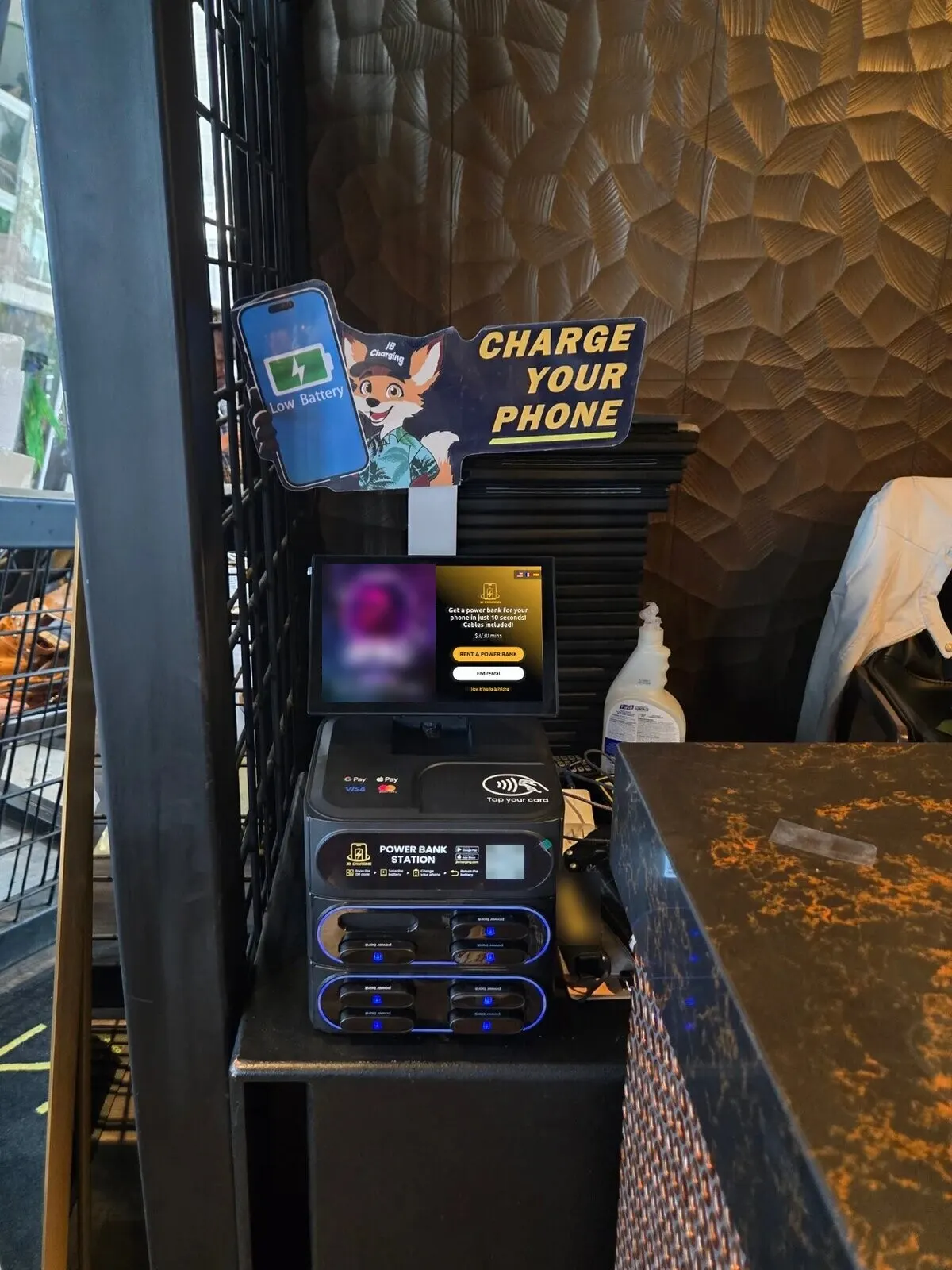 J8 power bank station with a "Charge Your Phone" mascot sign on a dark stand beside a dark counter, its screen showing a blurred ad next to a "Rent a power bank" button