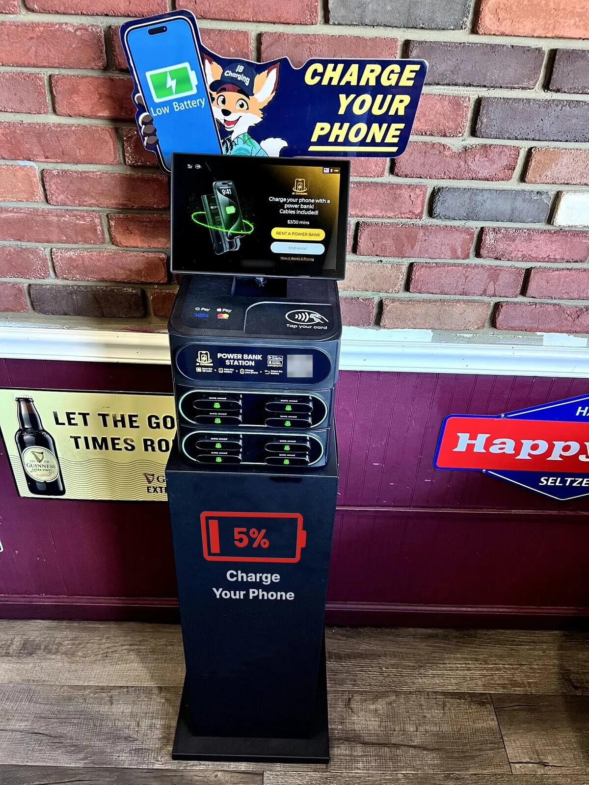 J8 power bank station on a black floor stand printed with a red 5% battery icon and "Charge Your Phone", under a fox-mascot "Charge Your Phone" header, against a red brick wall and maroon wainscoting in a pub, with beer signs on the wall and a wood floor.
