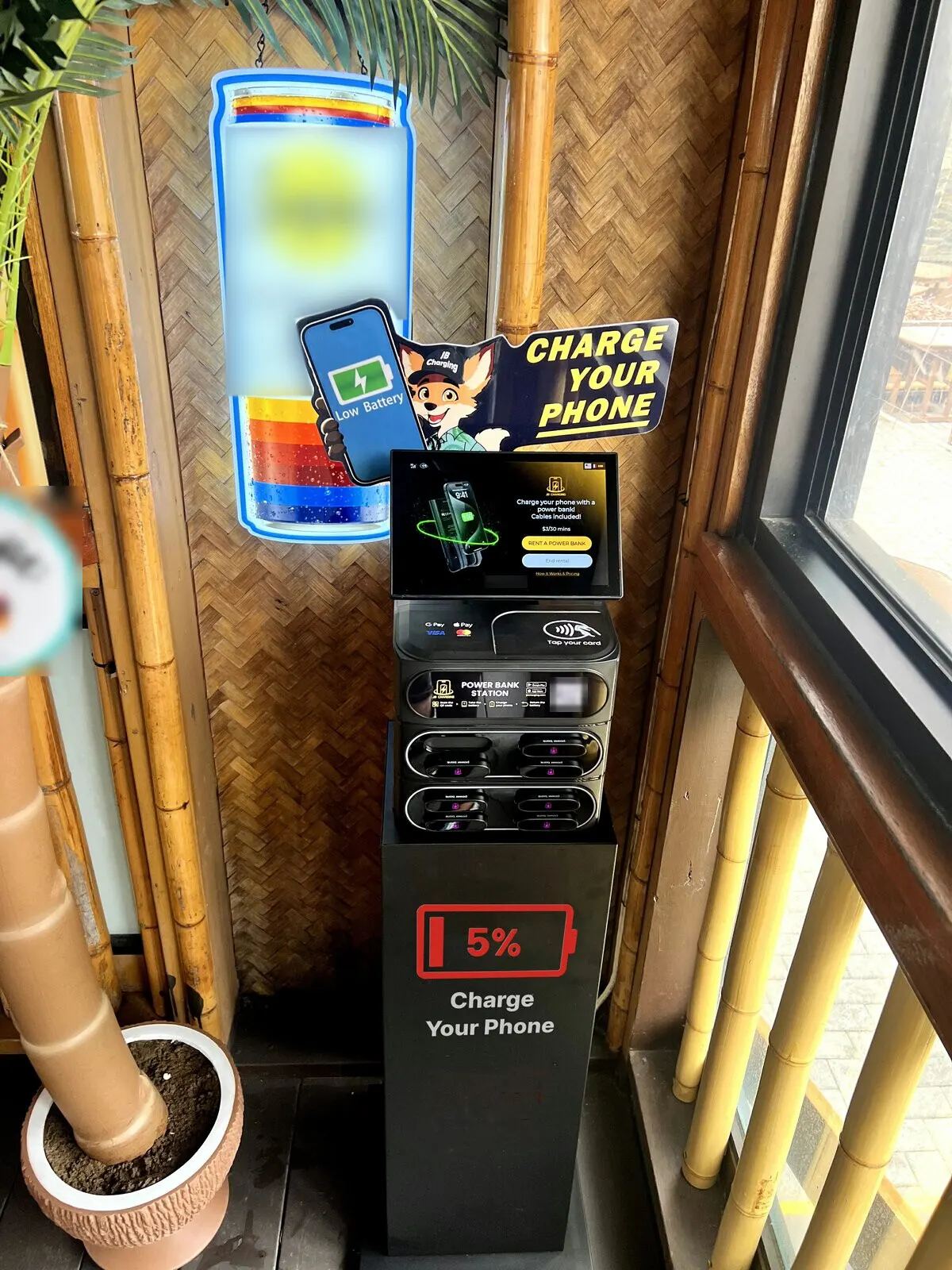 A JB Charging J8 Pro topped by a fox-mascot "Charge Your Phone" sign, on a black floor stand printed with a 5% battery icon, in a bamboo-walled corner of a bar beside a window and a potted palm
