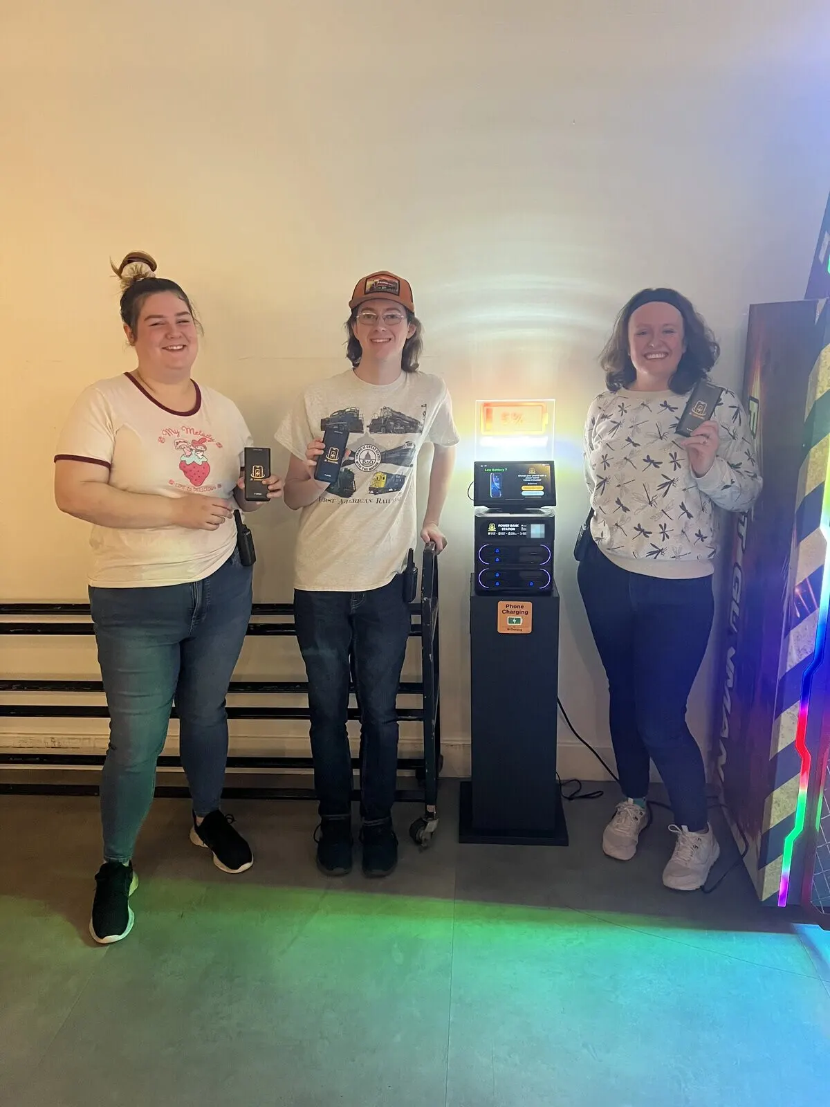 Three people holding JB Charging power banks beside a J8 station on a black stand, its lit header showing a 5% battery, with an arcade cabinet at the right edge