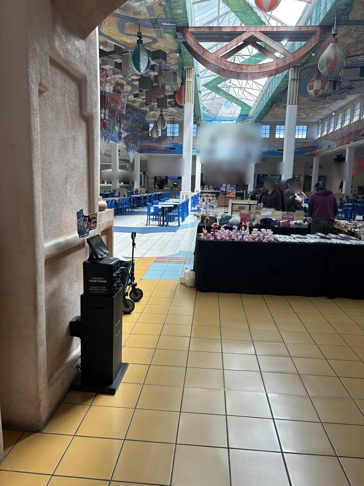 Black JB Charging countertop station on a black floor stand beside a stucco pillar, with blue food-court chairs behind it