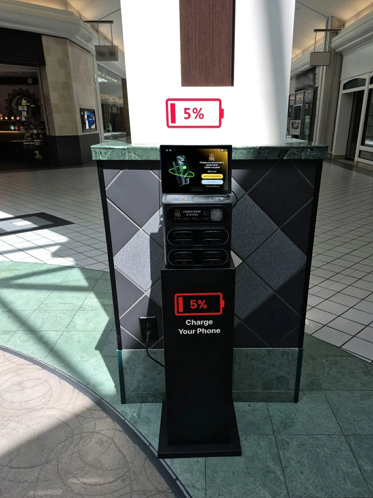 J8 power bank station on a black "5% Charge Your Phone" stand in a sunlit mall concourse, plugged into an outlet in the base of the pillar behind it, with a red 5% battery decal on the pillar above