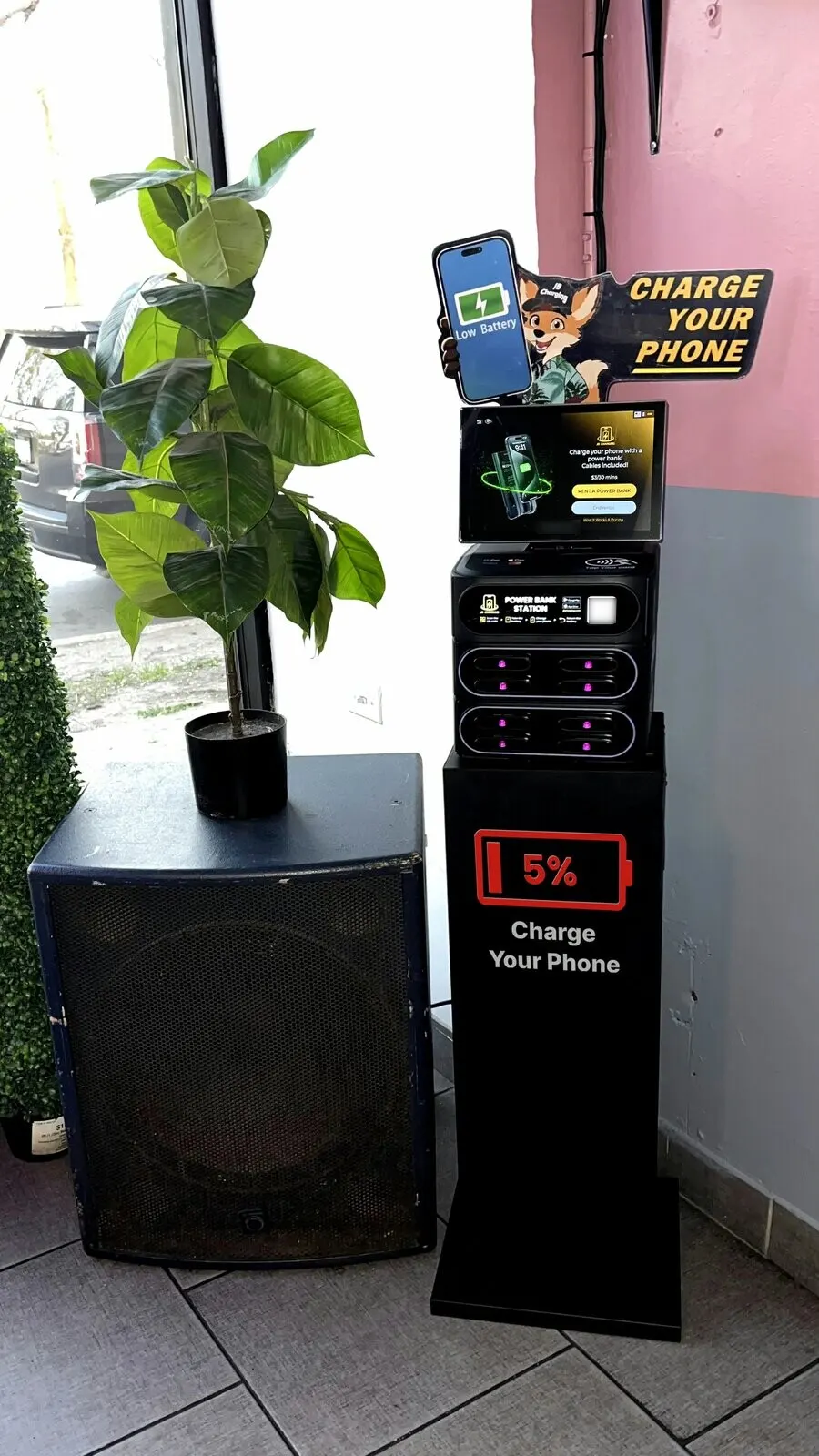J8 power bank station on a black "5% Charge Your Phone" floor stand, beside a PA speaker and a potted plant inside a front window