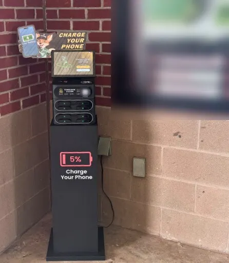A J8 power bank station with a cartoon "Charge Your Phone" sign on top, on a black stand in a brick and block corner, its cord running to a covered wall outlet beside a blurred field map