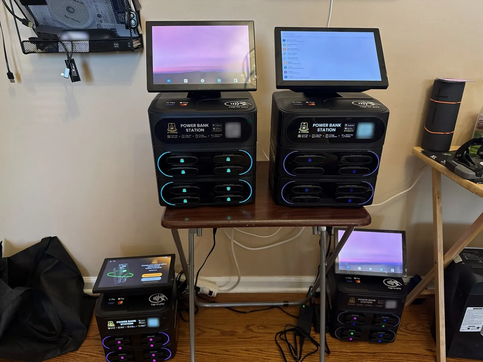 Four JB Charging countertop power bank stations powered on in a room at home, two on a small folding table and two on the floor beneath it, with home screens, a settings menu and one rental screen on their displays