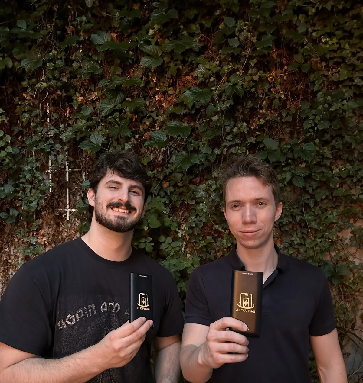 Two smiling men in dark shirts holding black JB Charging power banks toward the camera in front of an ivy-covered wall