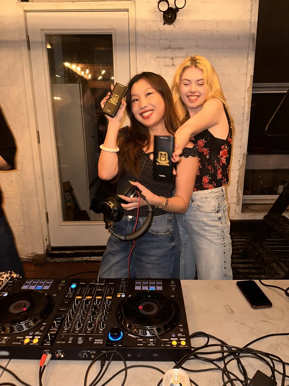 Two young women behind a DJ controller in front of a white brick wall, each holding up a black JB Charging power bank