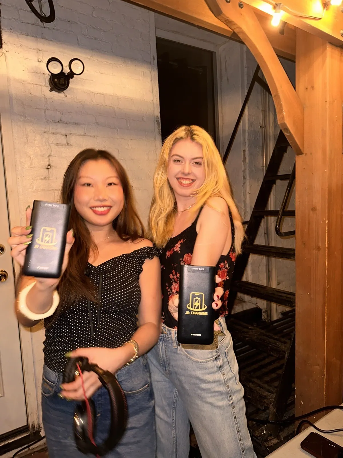 Two smiling women in front of a white brick wall and a black metal stair under string lights, each holding a black JB Charging power bank toward the camera; one holds headphones