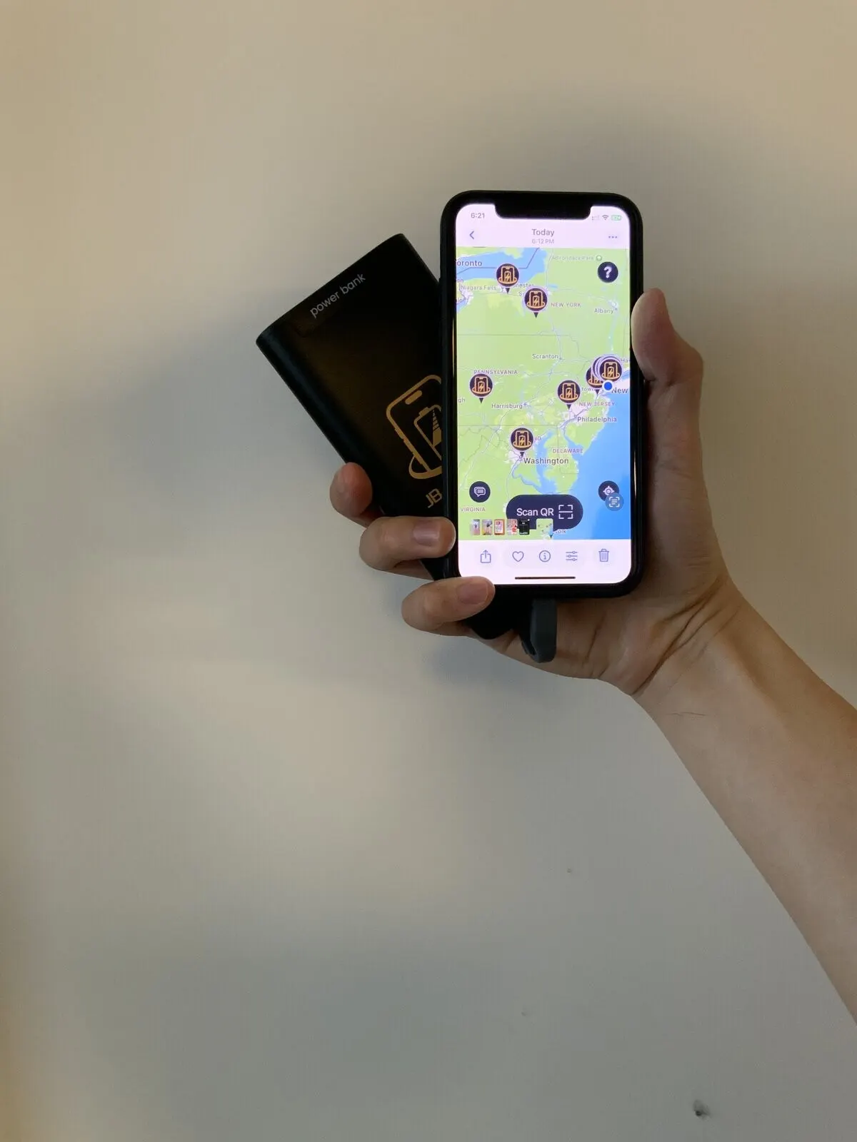 A hand holding an iPhone showing a map of JB Charging station pins across the Northeast with a Scan QR button, a black JB Charging power bank fanned behind it, against a plain beige wall