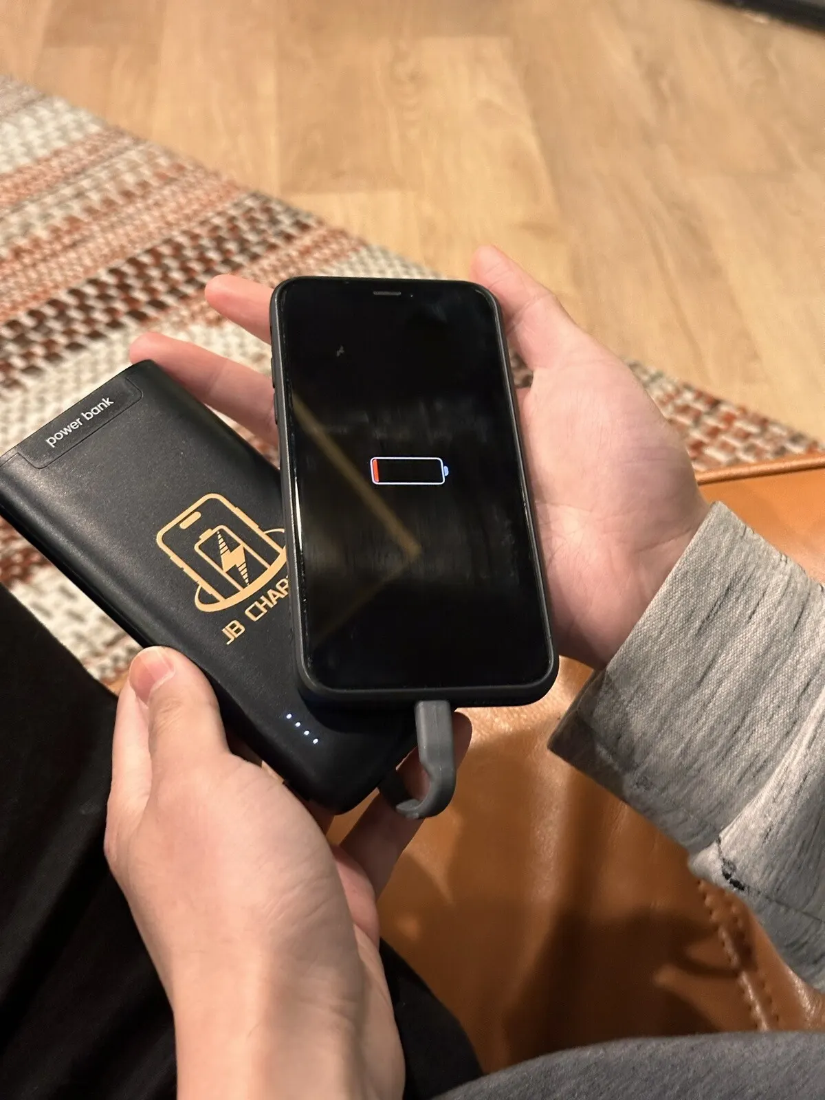 Hands holding an iPhone showing a nearly empty battery icon, plugged by a built-in cable into a black JB Charging power bank
