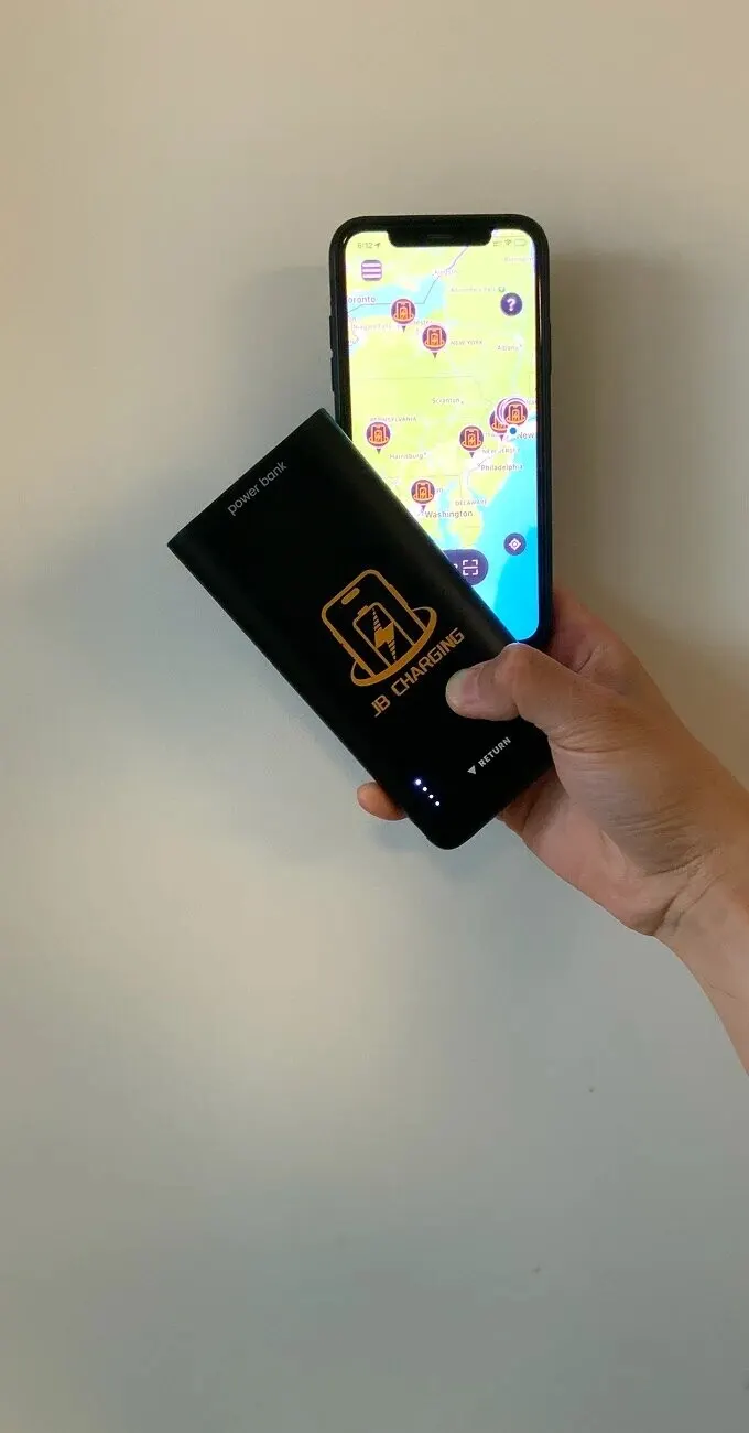 Hand holding a black JB Charging power bank in front of a phone showing a map of the Northeast US with station pins
