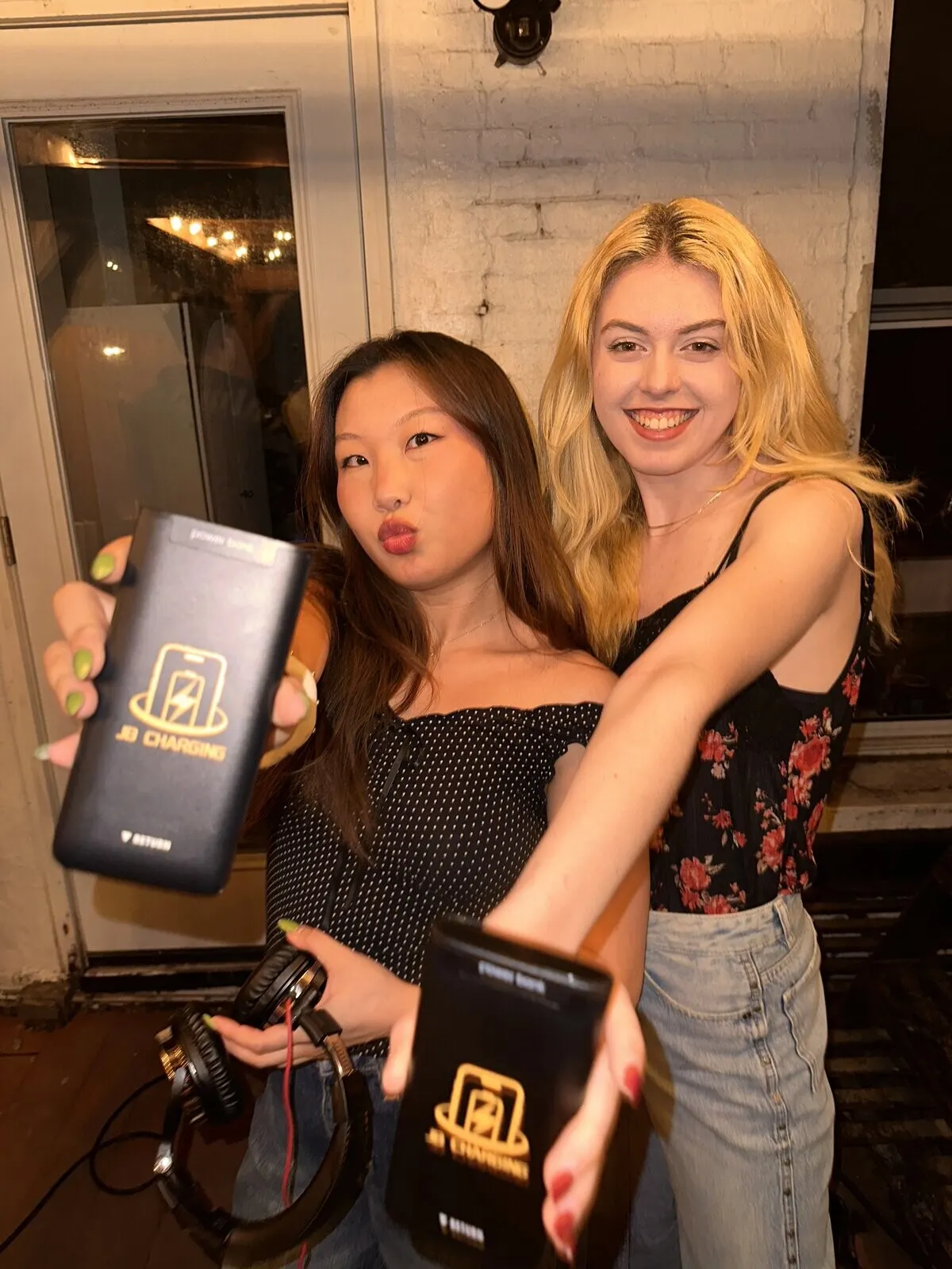 Two young women in front of a white brick wall at night, each holding a JB Charging power bank toward the camera, one also holding a pair of DJ headphones